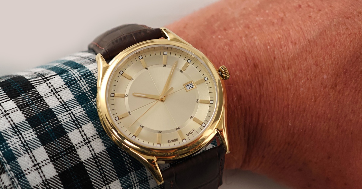 Close-up of the arm of a man wearing a plaid shirt showing a shiny gold watch with a thick leather strap.
